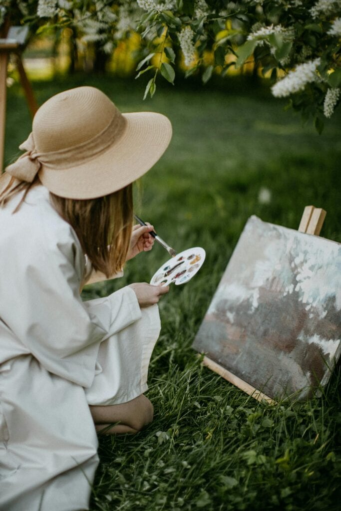 Characteristics Of Creativity: A Woman Sitting On The Grass And Painting On A Canvas.