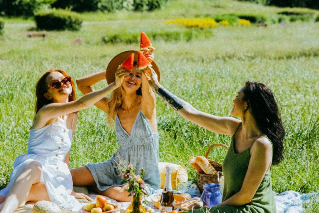 Three women on a picnic, cheering with a slice of watermelon in hand.