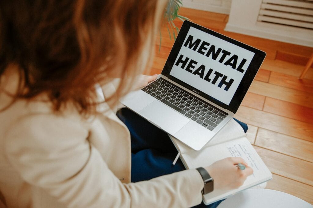 Woman seated on the floor, using a laptop, with text about mental health displayed on the screen.