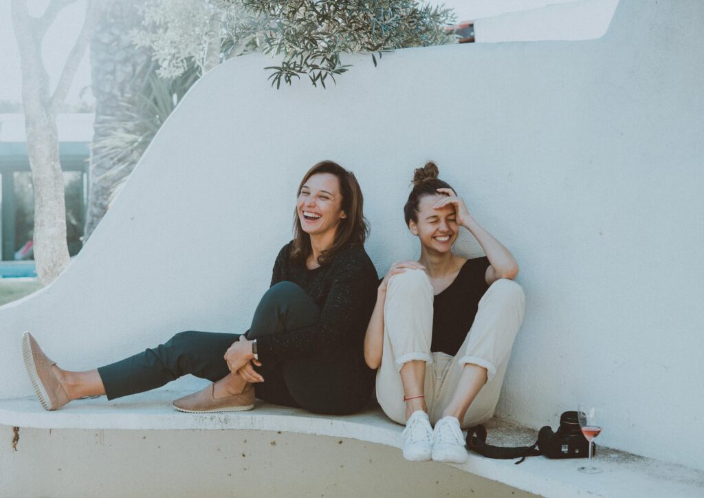 Two women sit on a wall, laughing and enjoying a sunny day together.