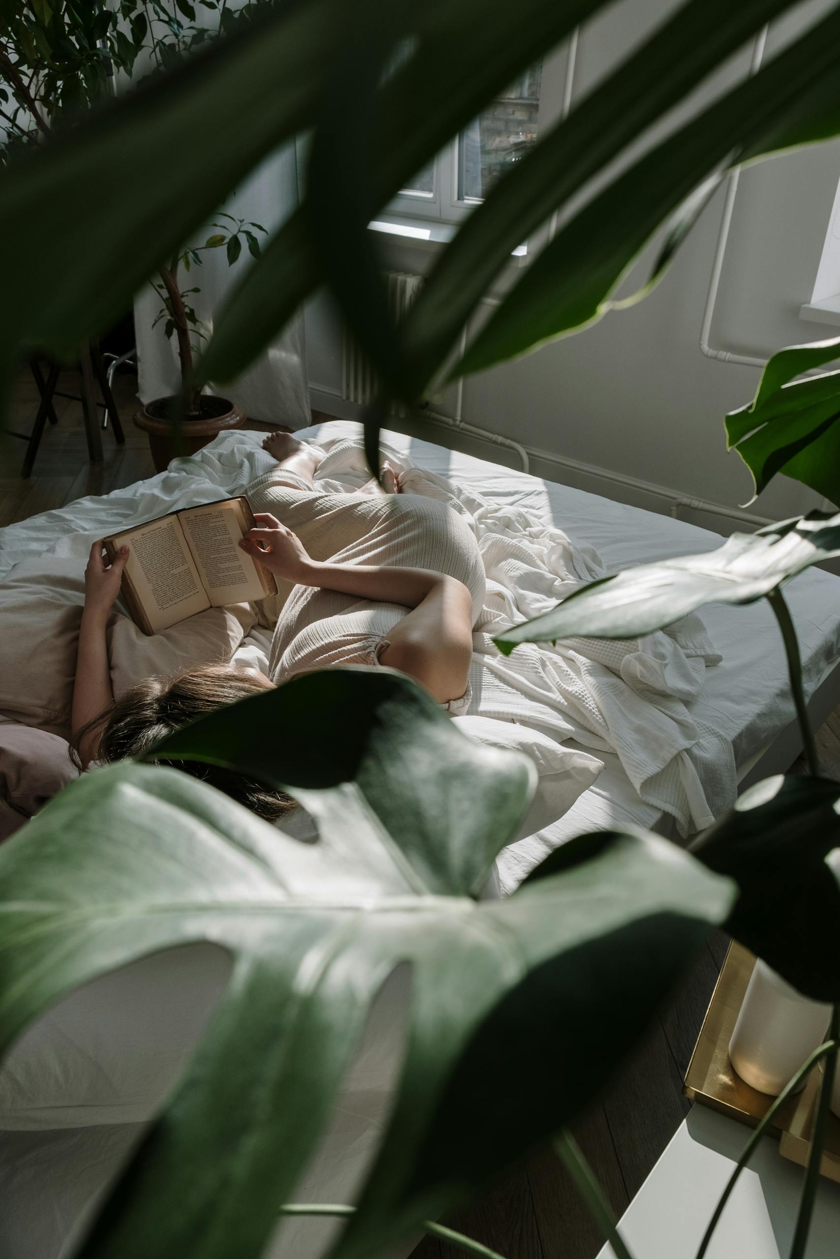 A woman lies in bed reading a book, surrounded by greenery and natural light. - How To Build A Great Nighttime Routine As Adults In 12 Steps