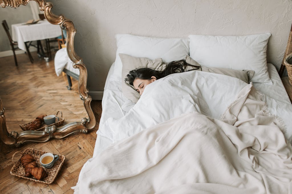 A woman peacefully sleeping in a cozy bed with croissants on the floor. - How To Build A Great Nighttime Routine As Adults In 12 Steps