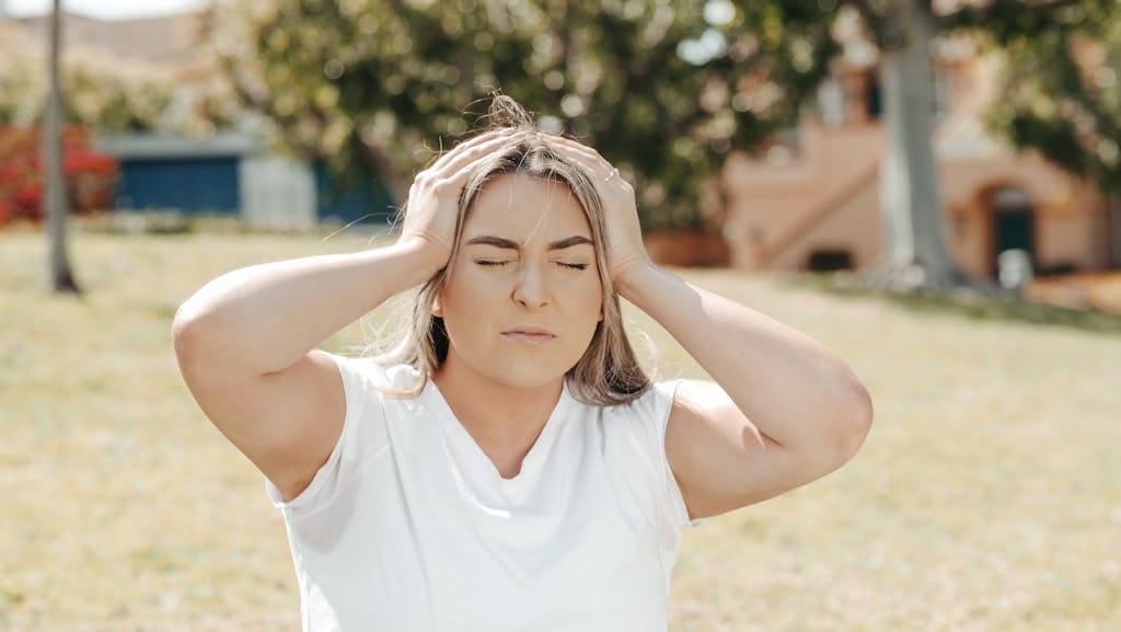 Adult woman holding her head in pain, sitting outdoors under sunlight, likely experiencing a migraine. - 11 Ways To Reprogram The Subconscious Mind For Success