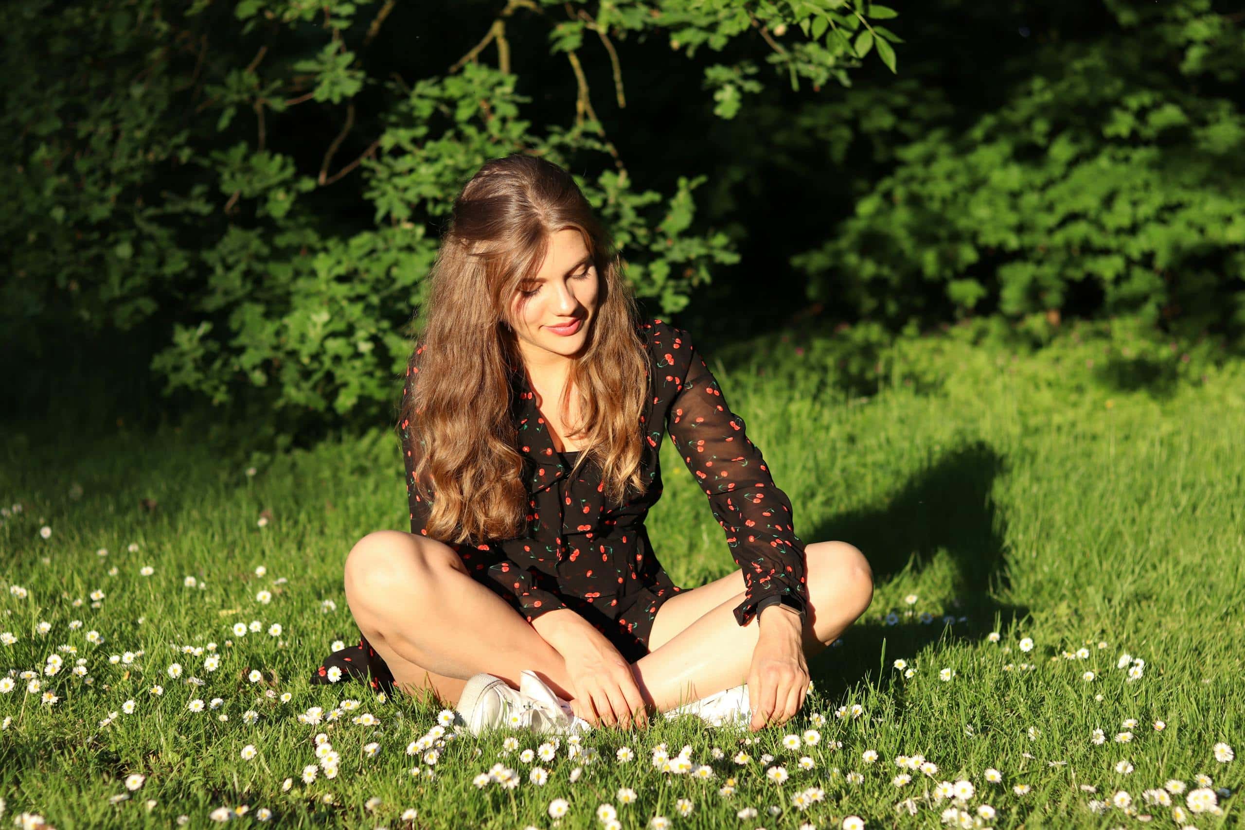 A woman in a black dress sits on grass in a sunlit field surrounded by daisies, exuding joy and relaxation. - How To Have A Healthy Mindset That Supports Growth