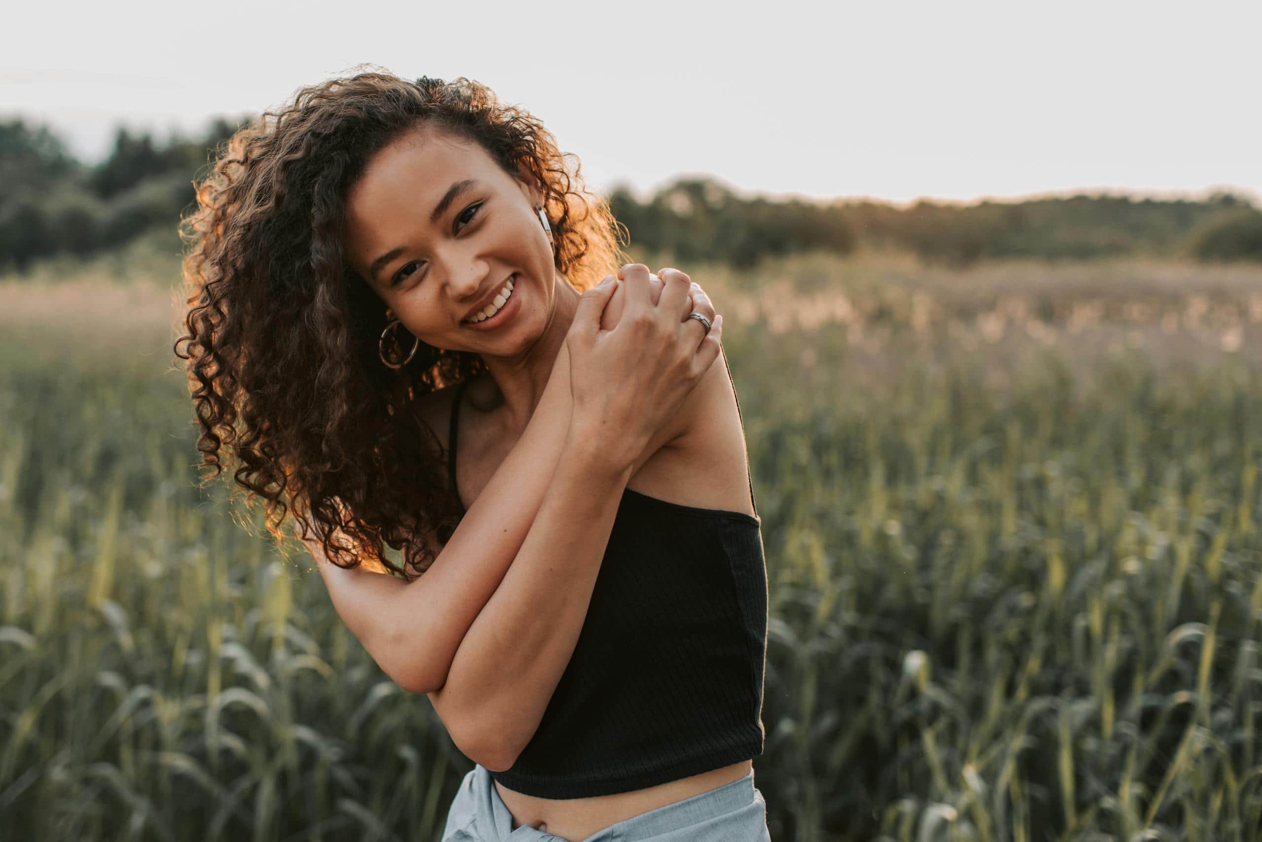 Joyful woman with curly hair smiling in a sunlit field, exuding carefree happiness. - How To Have A Healthy Mindset That Supports Growth