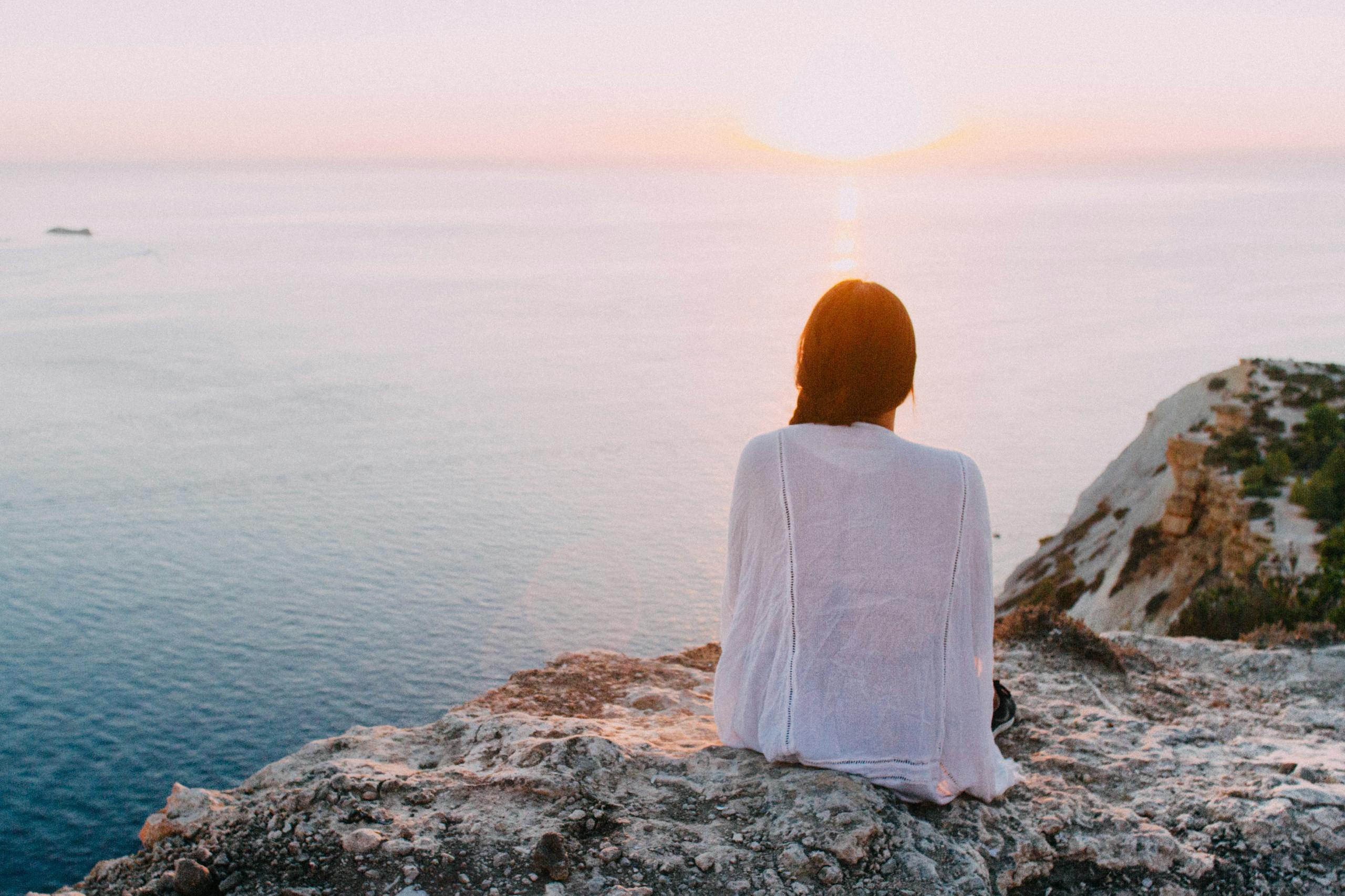A serene view of a woman sitting on a rocky cliff, gazing at the ocean during sunset. Perfect for relaxation and travel themes. | How I Started Living More Intentionally (You Can Too!)
