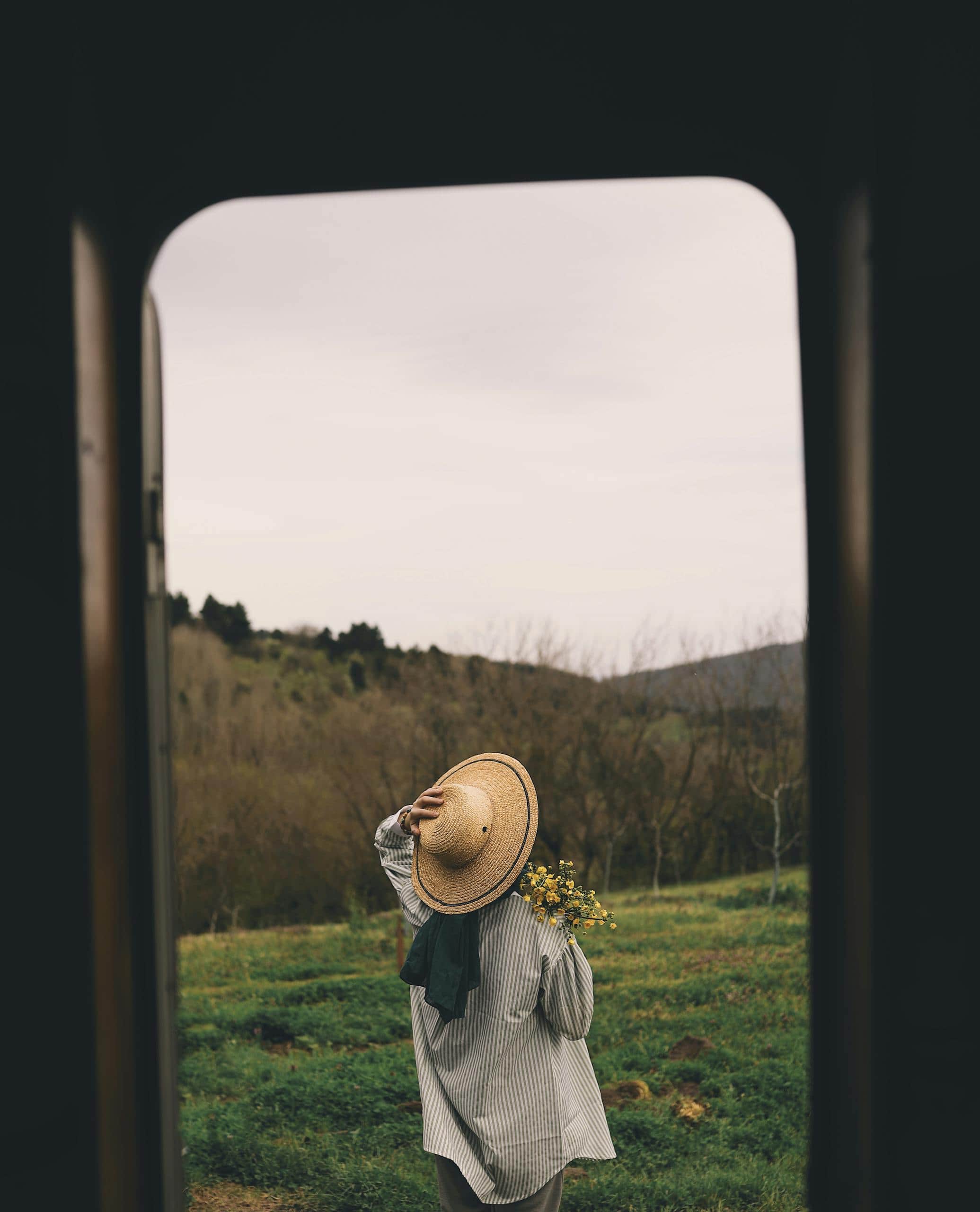 A woman wearing a wide-brimmed hat holds flowers while standing in a lush meadow viewed through a window frame. | How I Started Living More Intentionally (You Can Too!)