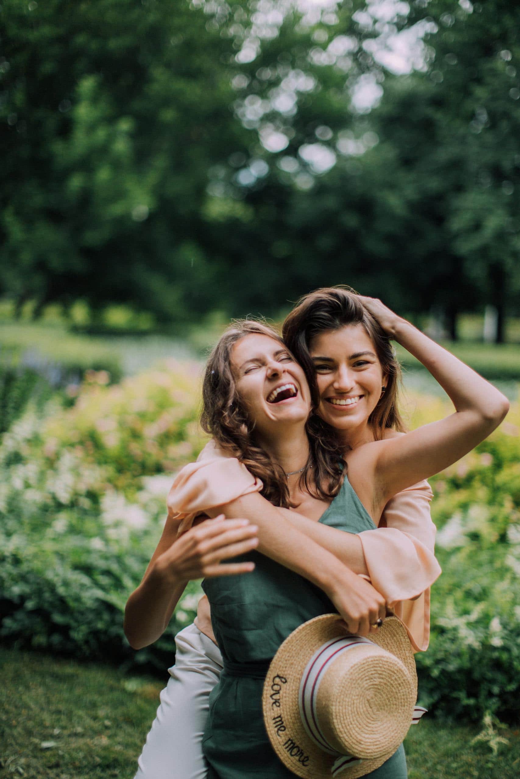 Two women laughing and hugging outdoors in a lush green park setting. Vibrant summer vibes. - How To Avoid Negative People: Surround Yourself With Positivity