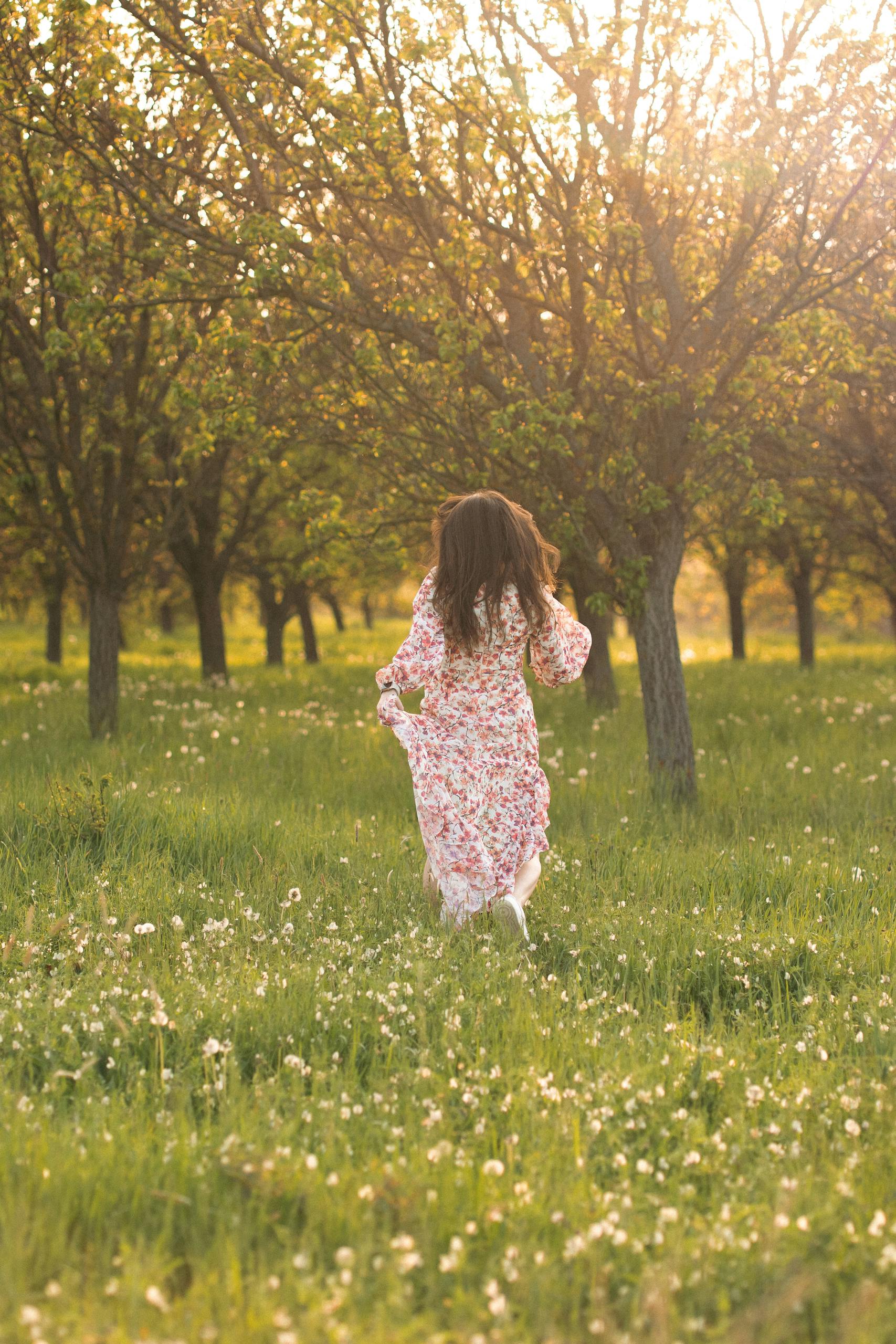 A young woman in a floral dress runs joyfully through a sunlit orchard, embracing nature. - How To Navigate A Life Transition Without Losing Yourself