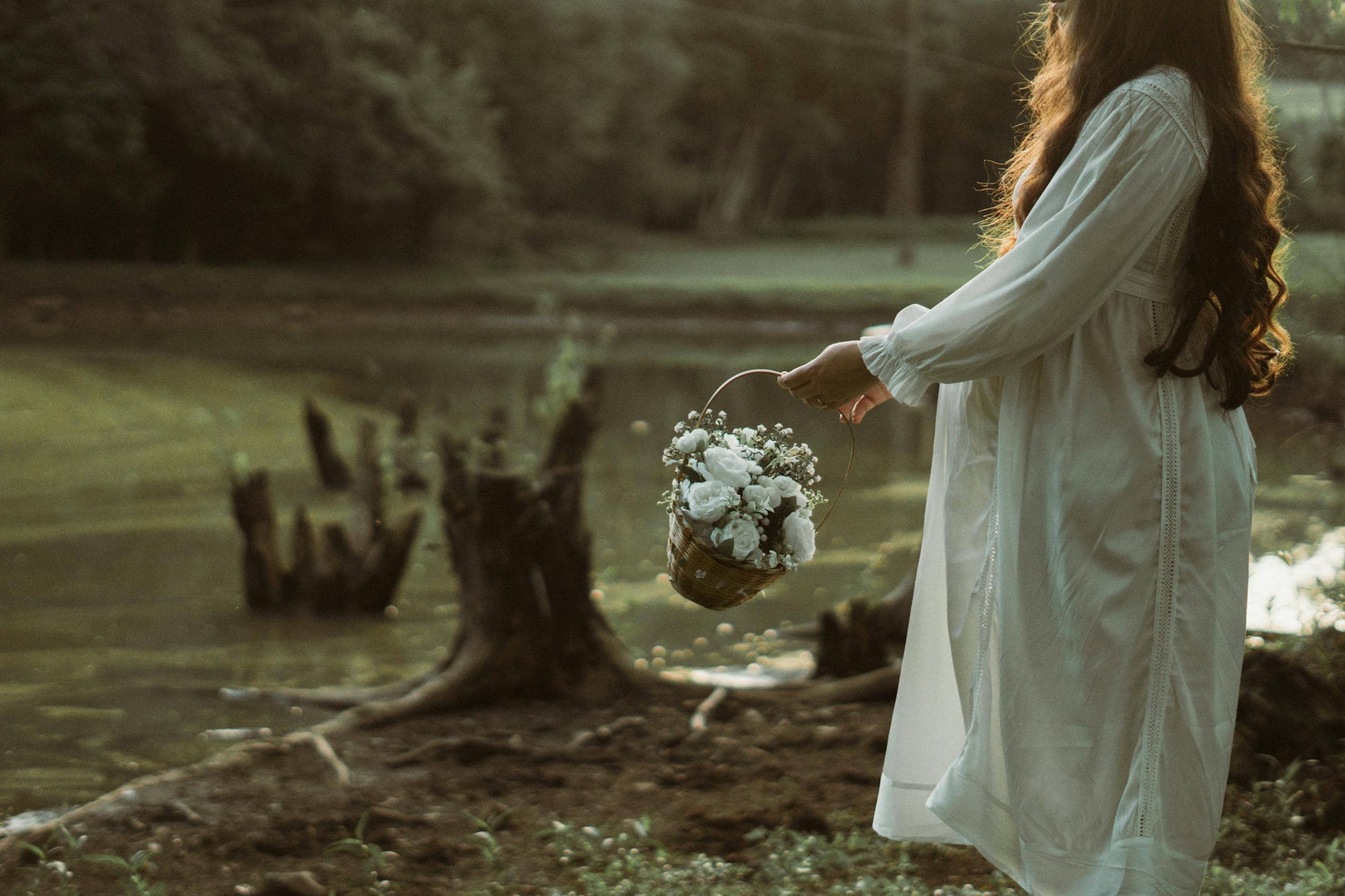 Pregnant woman in white dress holding a floral basket near a serene pond setting during the day. - How To Navigate A Life Transition Without Losing Yourself